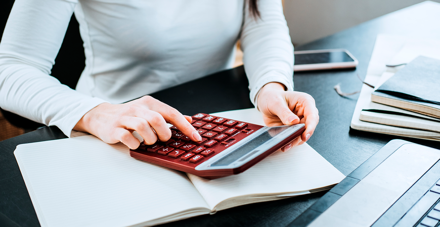Mulher usando calculadora vermelha sobre caderno aberto em mesa de escritório escura, celular e cadernos ao fundo, ambiente de análise financeira e contábil, luz natural suave, fotografia realista profissional